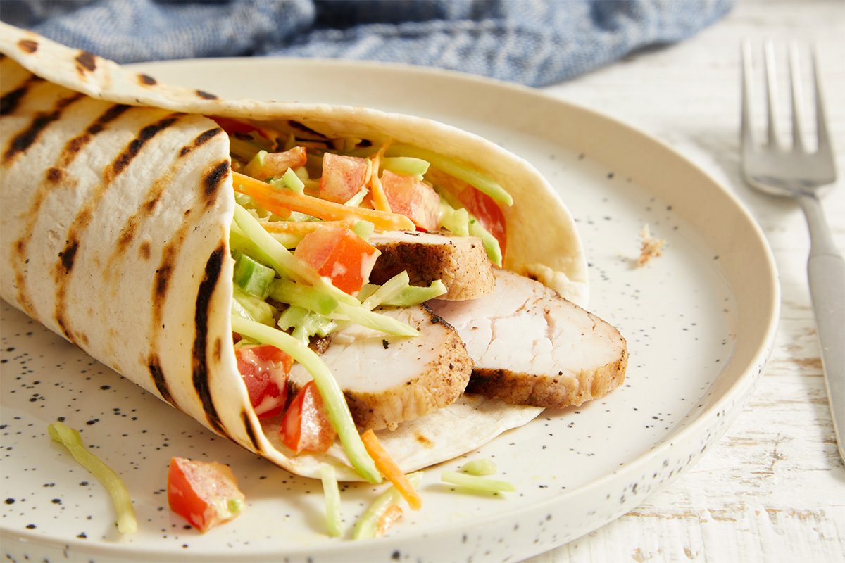 Eye level horizontal image of a Jamaican Jerk Turkey Wrap. The grilled flour tortilla is filled with sliced turkey tenderloin, chopped tomatoes, and a broccoli coleslaw mix. It is presented on a speckled plate with a fork and blue cloth in the background.