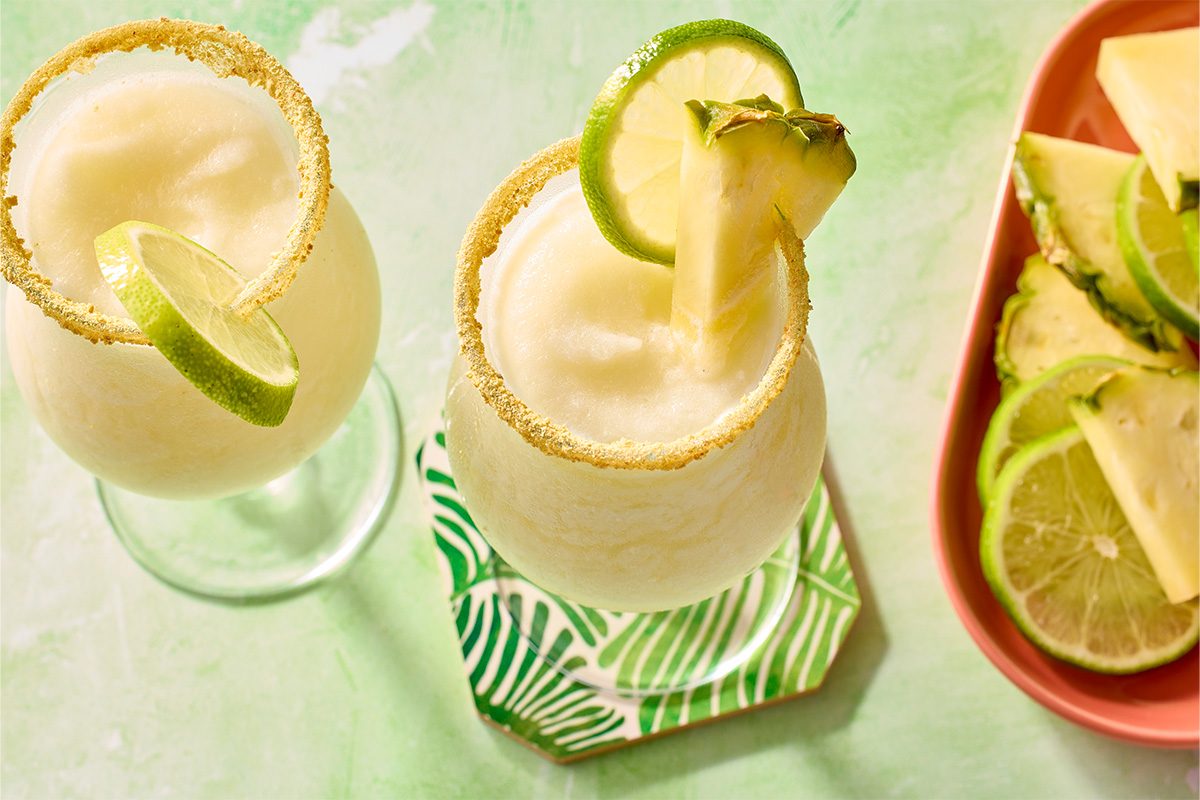 Overhead horizontal image of two frosted margarita glasses filled with a light-colored frozen beverage, likely a cocktail or smoothie. Both glasses have their rims coated in a sandy-colored crumb, and are garnished with fresh lime slices and pineapple wedges. A pink dish on the side holds additional lime slices and pineapple wedges.