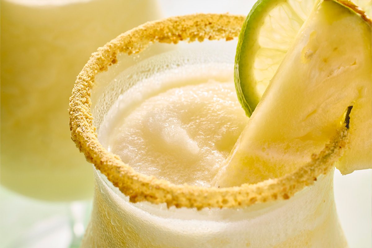 Eye Level horizontal image of a frosty glass filled with a light yellow, blended frozen drink. The rim of the glass is coated with golden crumbs, and the drink is garnished with a fresh lime slice and a wedge of pineapple.