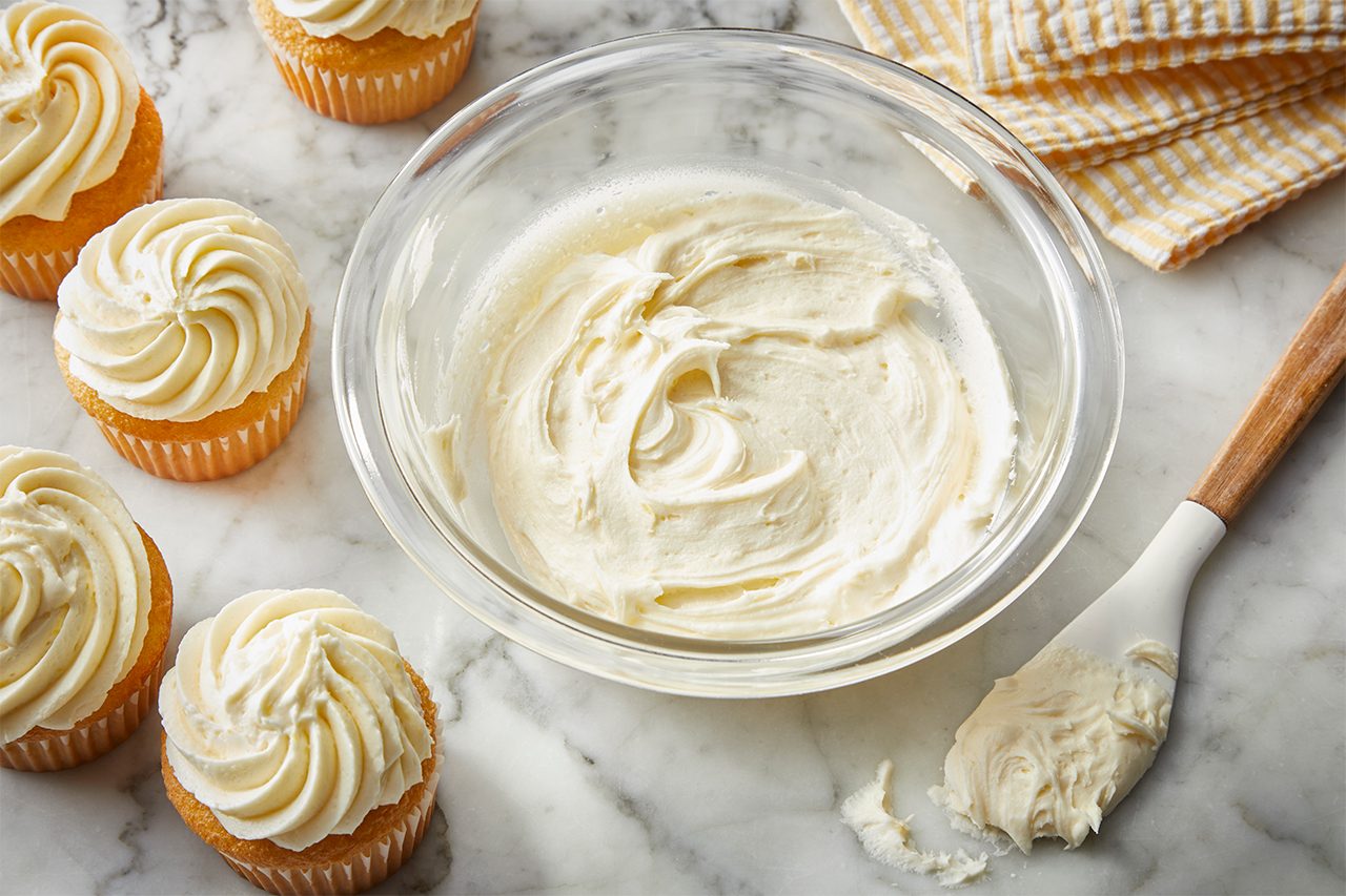 A glass bowl of creamy white frosting sits on a marble surface with a spatula beside it. Five vanilla cupcakes topped with swirled frosting are arranged nearby. A folded yellow checkered cloth is in the background.