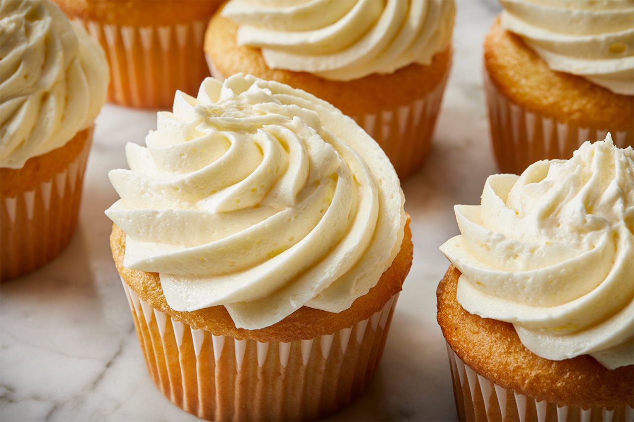Vanilla cupcakes with swirls of creamy white frosting are arranged on a marble surface.