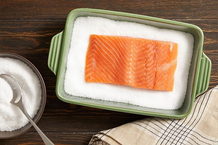 Overhead horizontal image of a salmon fillet being prepared on a bed of coarse white salt in a green rectangular baking dish. To the left, a bowl filled with more salt and a silver spoon is visible on a dark wooden surface, alongside a checkered napkin.