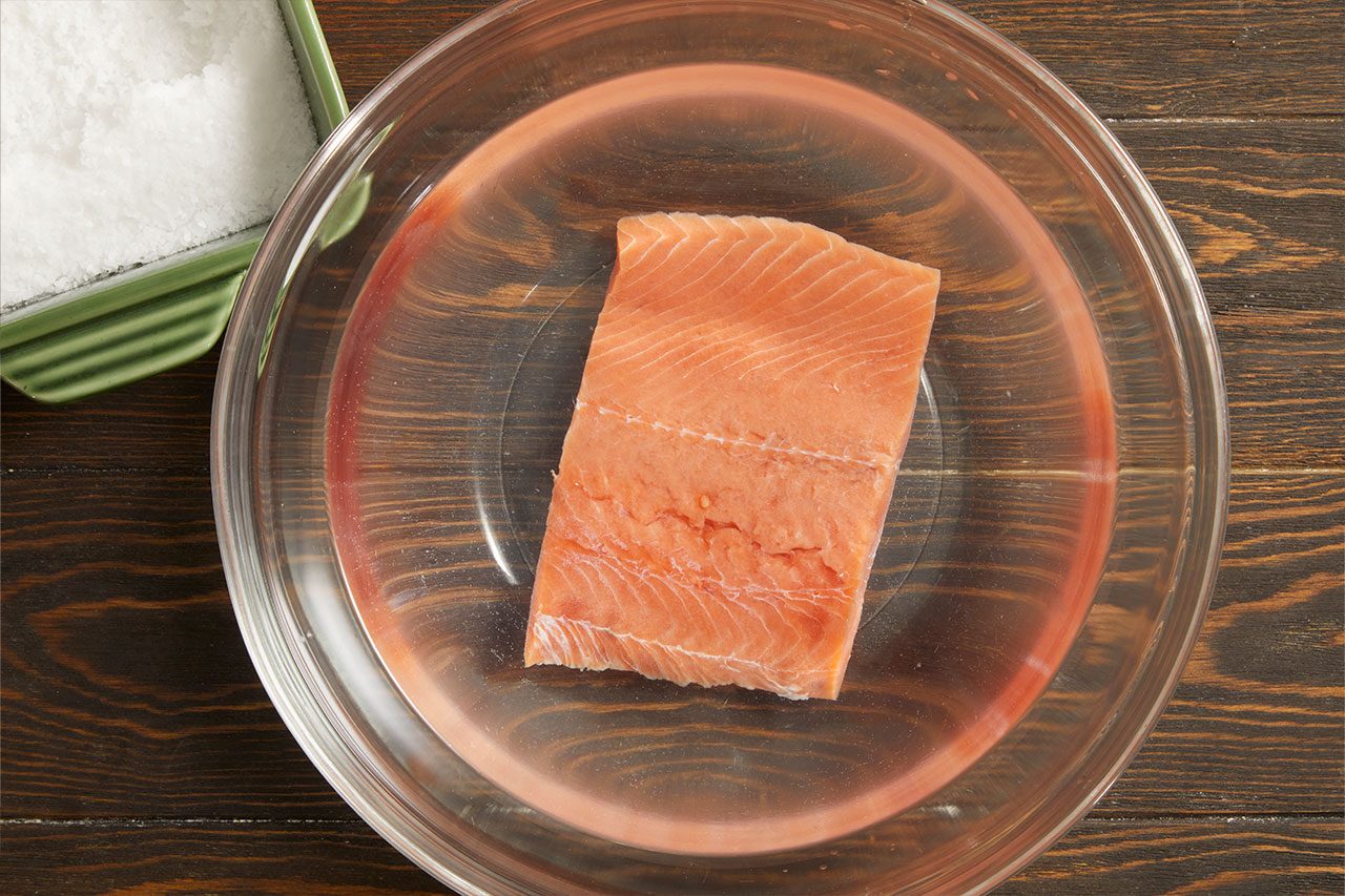 Overhead horizontal image of a raw salmon fillet soaking in a clear liquid within a glass bowl. A green dish filled with white granular salt is partially visible in the top left corner, suggesting a brining or curing process.