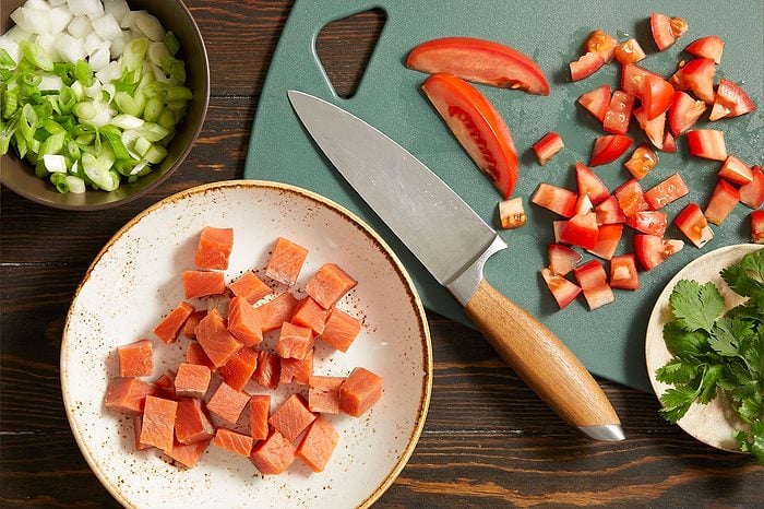 Overhead horizontal image of various fresh ingredients being prepped on a dark wooden surface. Cubed salmon is on a white plate next to a green cutting board with diced tomatoes and a chef's knife. A bowl of diced white onion and chopped green onions, along with a small plate of fresh cilantro, are also visible.