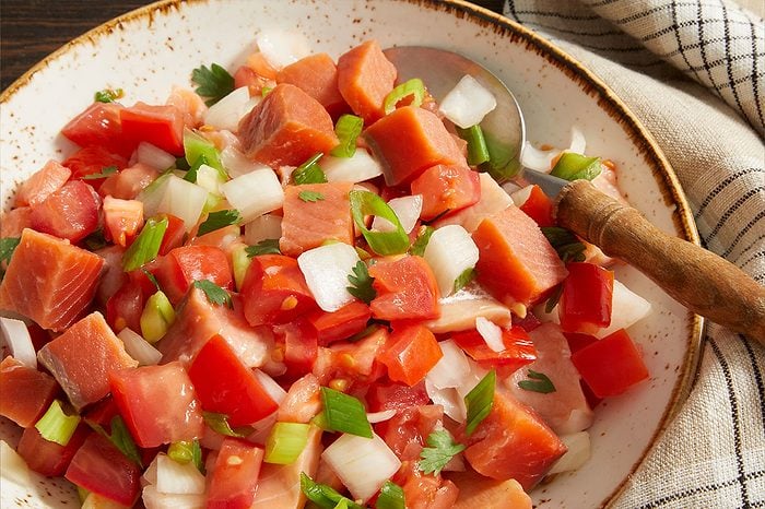 Overhead horizontal image of a white speckled bowl filled with a vibrant mix of ingredients. The dish features diced red fish, likely salmon, combined with bright red diced tomatoes, white onions, and chopped green onions, garnished with fresh parsley. A silver spoon with a wooden handle rests in the food.