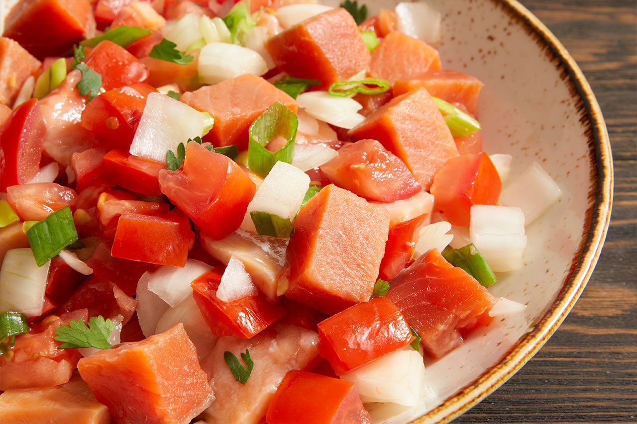 Eye Level horizontal image of a close-up on a bowl filled with a vibrant mixture of diced ingredients. The dish features chunks of reddish-orange fish, bright red tomatoes, white onion, and sliced green onions, garnished with fresh cilantro. It is presented in a ceramic bowl with a rustic speckled rim, set against a dark wooden background.