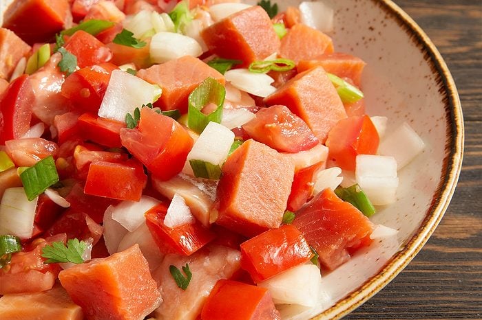 Eye Level horizontal image of a close-up on a bowl filled with a vibrant mixture of diced ingredients. The dish features chunks of reddish-orange fish, bright red tomatoes, white onion, and sliced green onions, garnished with fresh cilantro. It is presented in a ceramic bowl with a rustic speckled rim, set against a dark wooden background.