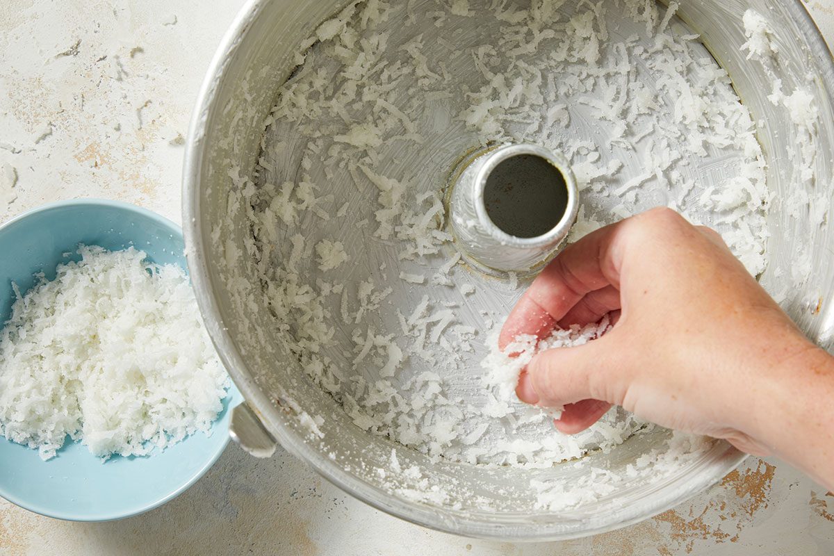 Overhead horizontal image of a hand sprinkling shredded coconut into a greased bundt pan. A light blue bowl filled with shredded coconut is visible to the left, ready for use.
