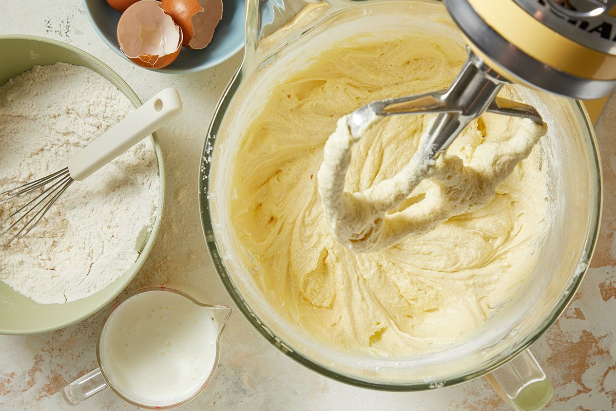 Overhead horizontal image of a stand mixer actively beating a light yellow batter in a glass bowl. To the left, a bowl of white flour with a whisk rests next to a measuring cup filled with milk. In the background, a small bowl holds brown eggshells, indicating eggs were recently added to the mixture.