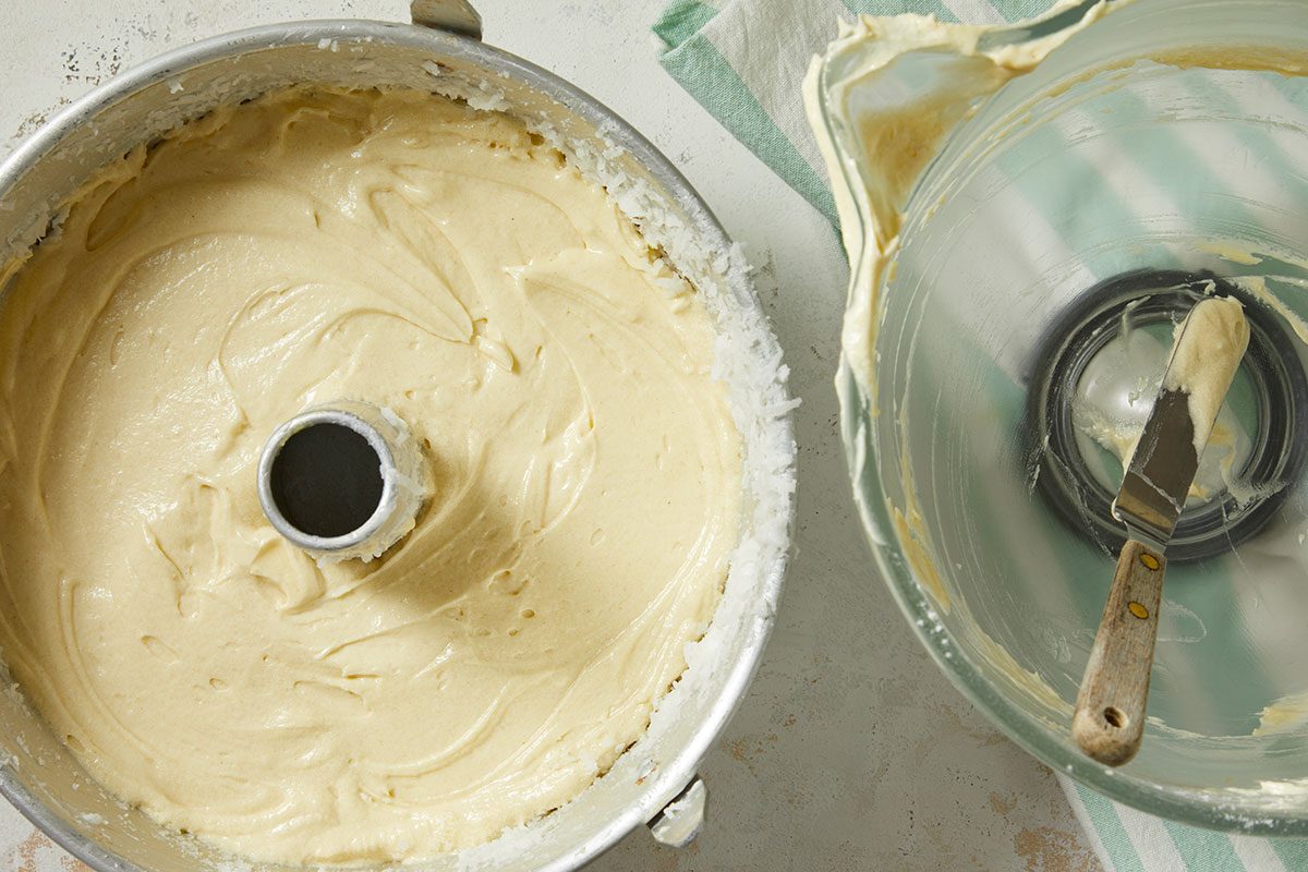 Overhead horizontal image of light yellow cake batter filling a fluted bundt pan, which appears to be coated with shredded coconut. Next to it, a clear glass mixing bowl with residual batter and a spatula rests on a striped kitchen towel.