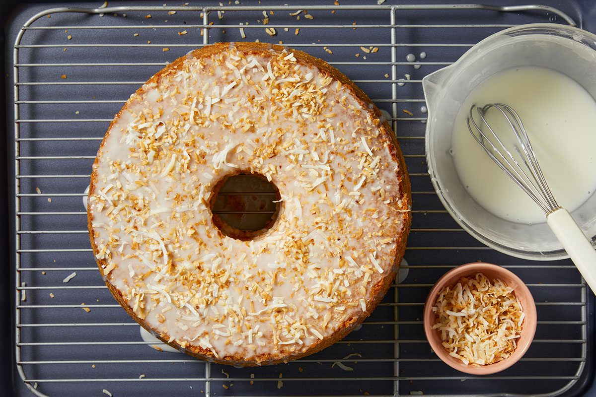 Overhead horizontal image of a freshly glazed bundt cake, generously topped with toasted coconut, resting on a metal cooling rack. Alongside the cake, a clear measuring cup contains additional white glaze with a whisk, and a small pink bowl holds more toasted coconut flakes.