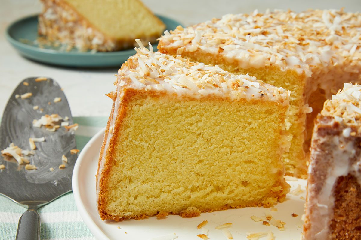 Eye Level horizontal image of a slice of cake being served from a large cake. The golden-brown cake and slice are generously topped with white glaze and toasted coconut flakes, with a serving utensil on the side.