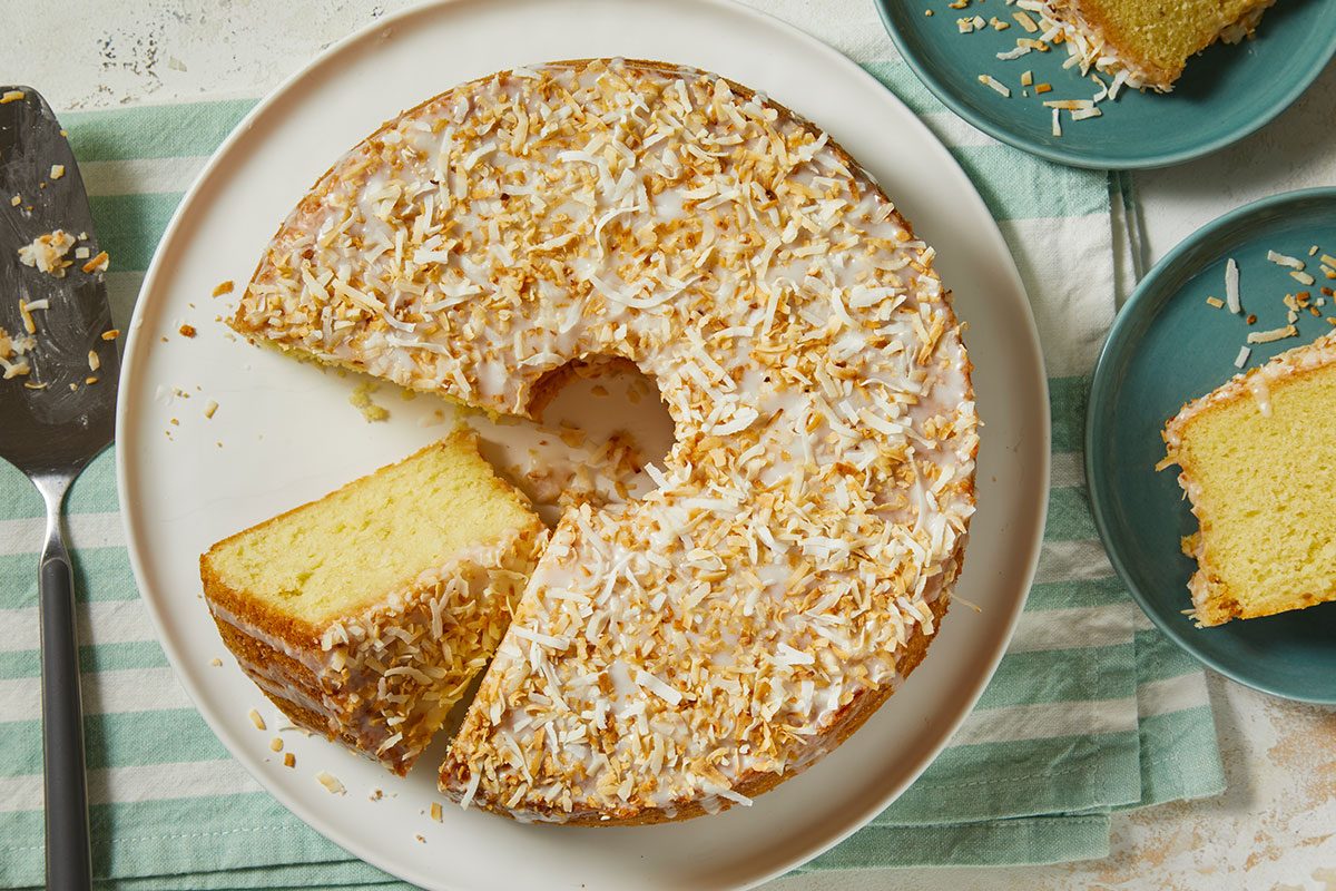 Overhead horizontal image of a round cake with a slice removed, topped with a white glaze and toasted coconut flakes. Two additional slices are on small blue plates next to the main cake, which rests on a striped napkin with a cake server alongside.
