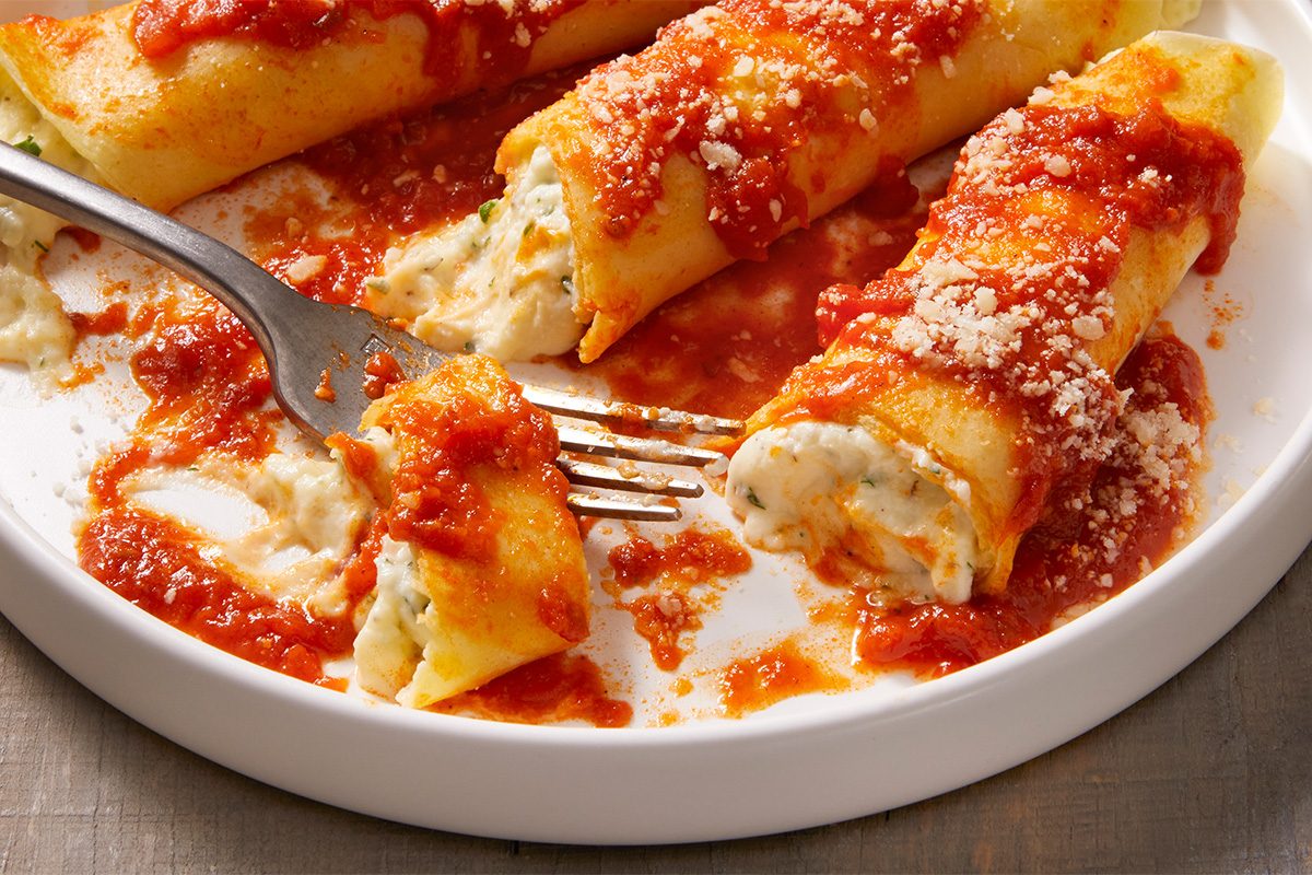Overhead close-up food photography of Homemade Manicotti being cut with a fork, revealing creamy ricotta filling inside crepe noodles.