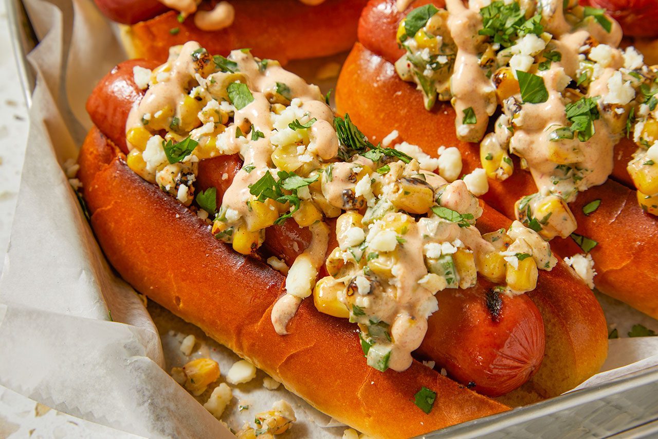 Overhead shot of Mexican Street Corn Hot Dogs arranged on a parchment-lined baking tray, topped with a creamy grilled corn mixture, crumbled cotija cheese, chopped cilantro, and a drizzle of sauce. Lime wedges, fresh cilantro, and a striped kitchen towel are arranged nearby on a light countertop.