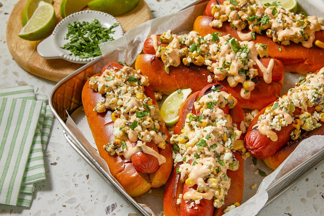 Close-up shot of a Mexican Street Corn Hot Dog topped with a creamy grilled corn mixture, crumbled cotija cheese, chopped cilantro, and a drizzle of sauce, highlighting the loaded toppings and toasted bun, with additional hot dogs softly blurred in the background.