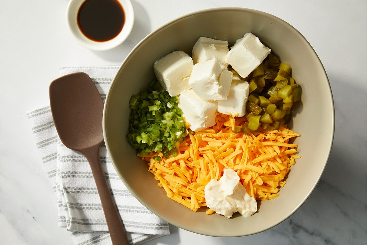 Overhead horizontal image of a gray bowl filled with cubed cream cheese, chopped dill pickles, chopped green onions, shredded cheddar cheese, and a dollop of mayonnaise. A brown silicone spatula rests on a striped white kitchen towel, and a small white dish containing Worcestershire sauce is visible nearby. | [Original] RMS 38691 Mini Dill Pickle Cheese Balls; How to; Step 1; ingredients for cheese ball in bowl, not yet combined
