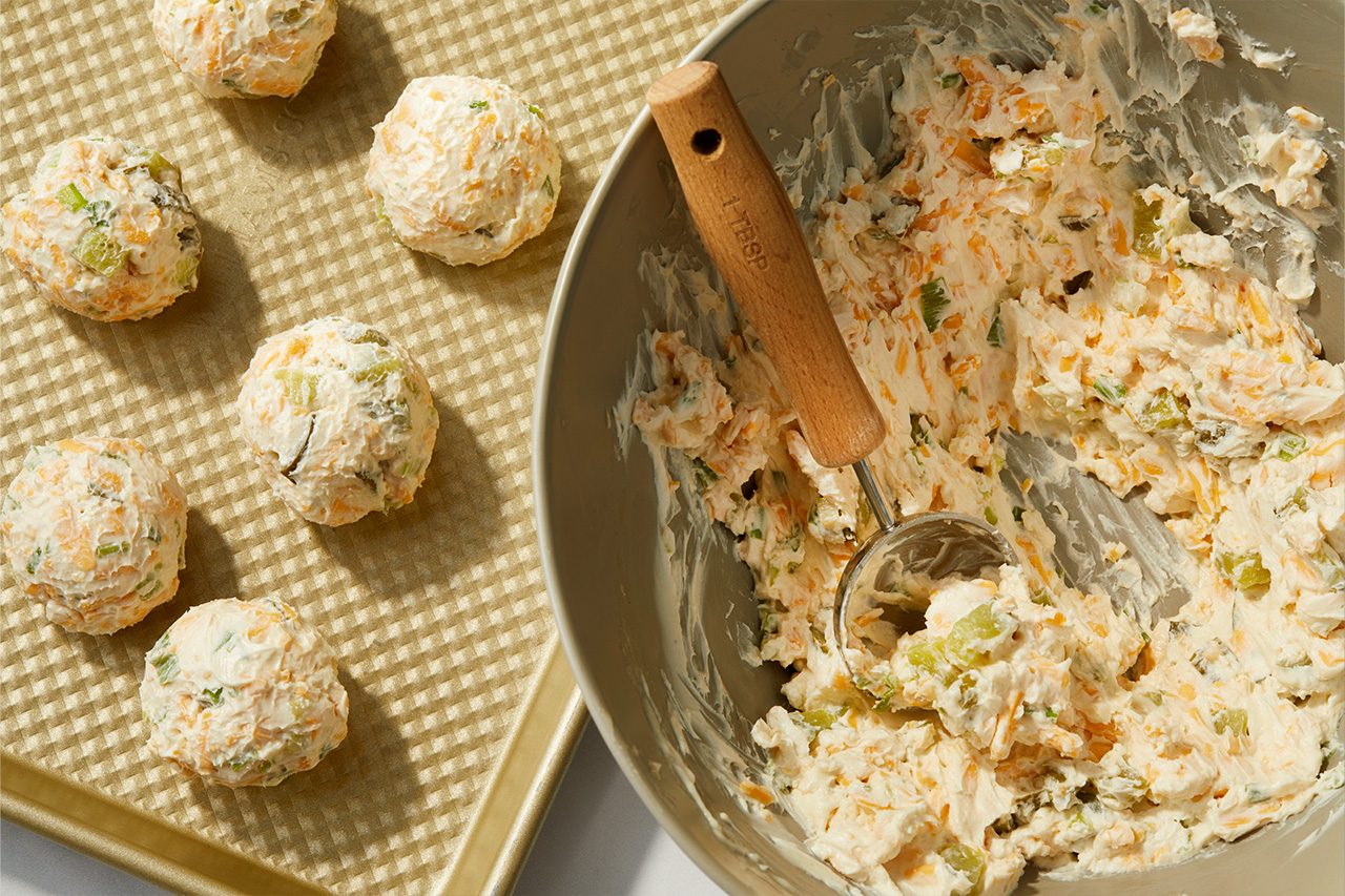 Overhead horizontal image of mini cheese balls being formed. Several creamy, orange, and green-speckled cheese balls are arranged on a gold baking sheet. On the right, a gray bowl holds the remaining mixture, which includes softened cream cheese, shredded cheddar, chopped dill pickles, and green onions, alongside an ice cream scoop. | [Original] RMS 38691 Mini Dill Pickle Cheese Balls; How to; Step 2; Rolling cheese mixture into balls