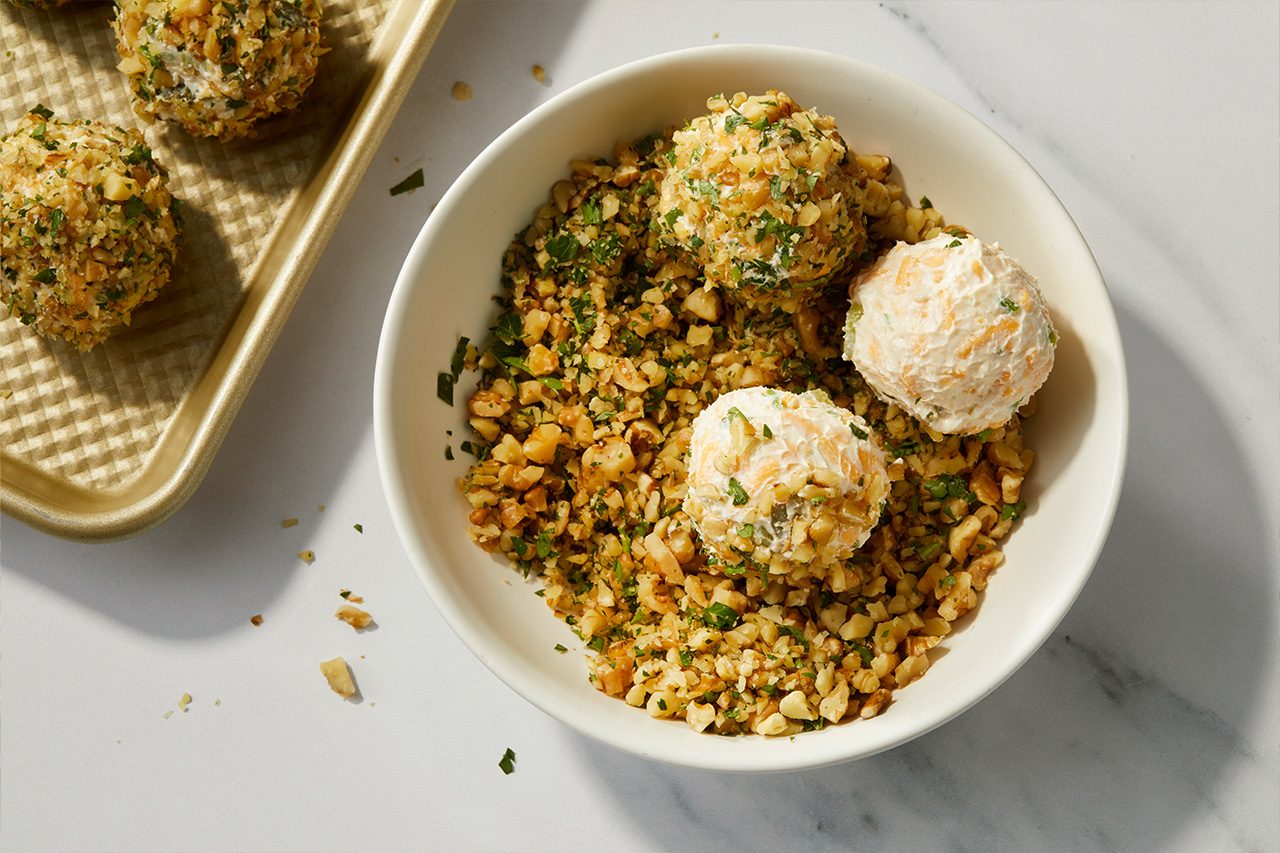 Overhead horizontal image of several Dilly Mini Cheese Balls being coated. A white bowl holds a mixture of chopped walnuts and minced fresh parsley, where the creamy, cheesy balls are rolled. Some cheese balls are fully coated, while others are partially visible, revealing their rich texture. | [Original] RMS 38691 Mini Dill Pickle Cheese Balls; How to; Step 3; Coating cheese balls in walnut mixture