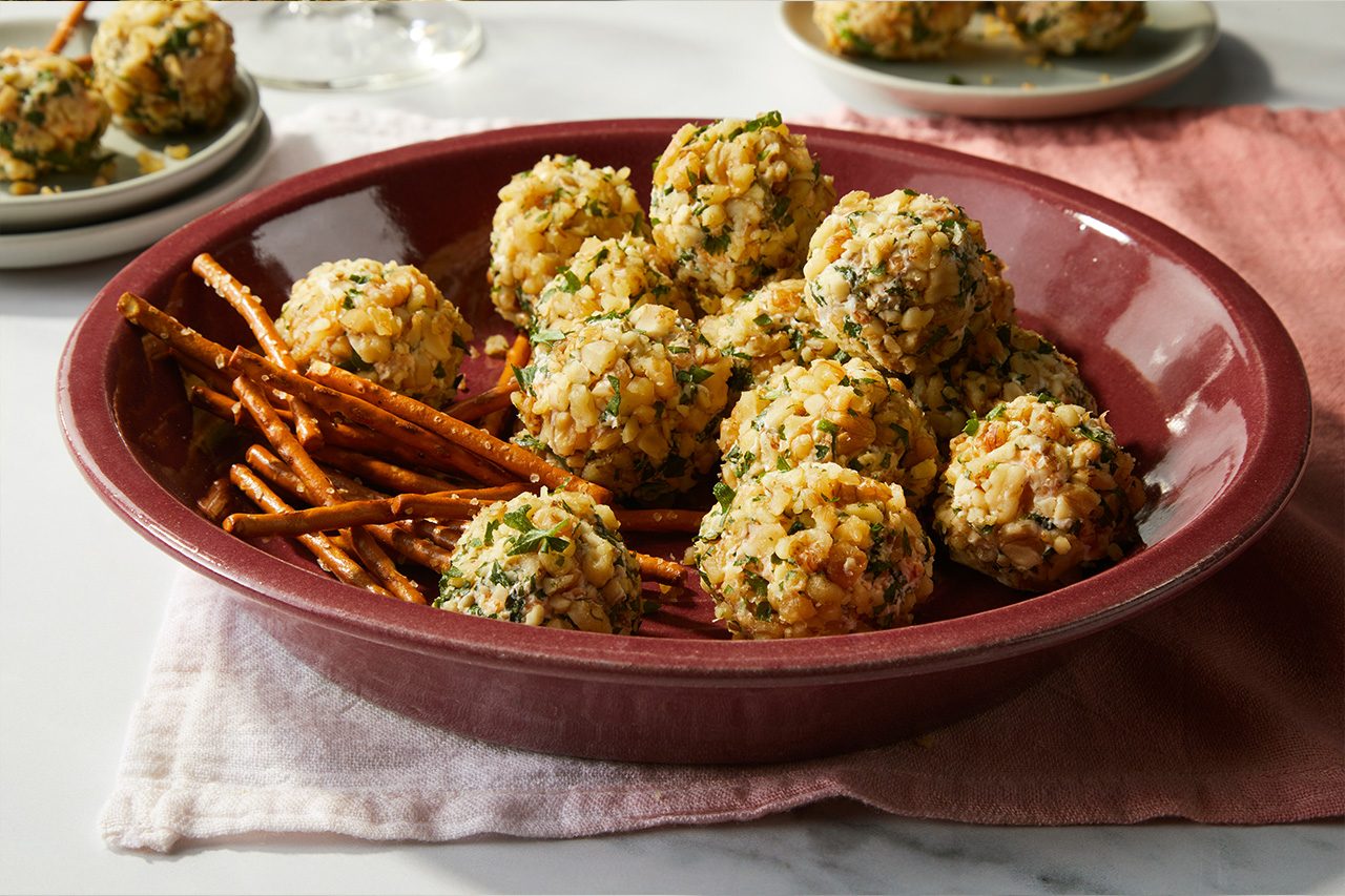 Eye Level horizontal image of mini cheese balls and pretzel sticks served in a maroon bowl on a marble surface. The cheese balls are coated in chopped walnuts and fresh parsley. | [Original] RMS 38691 Mini Dill Pickle Cheese Balls; Horizontal AP; Pull