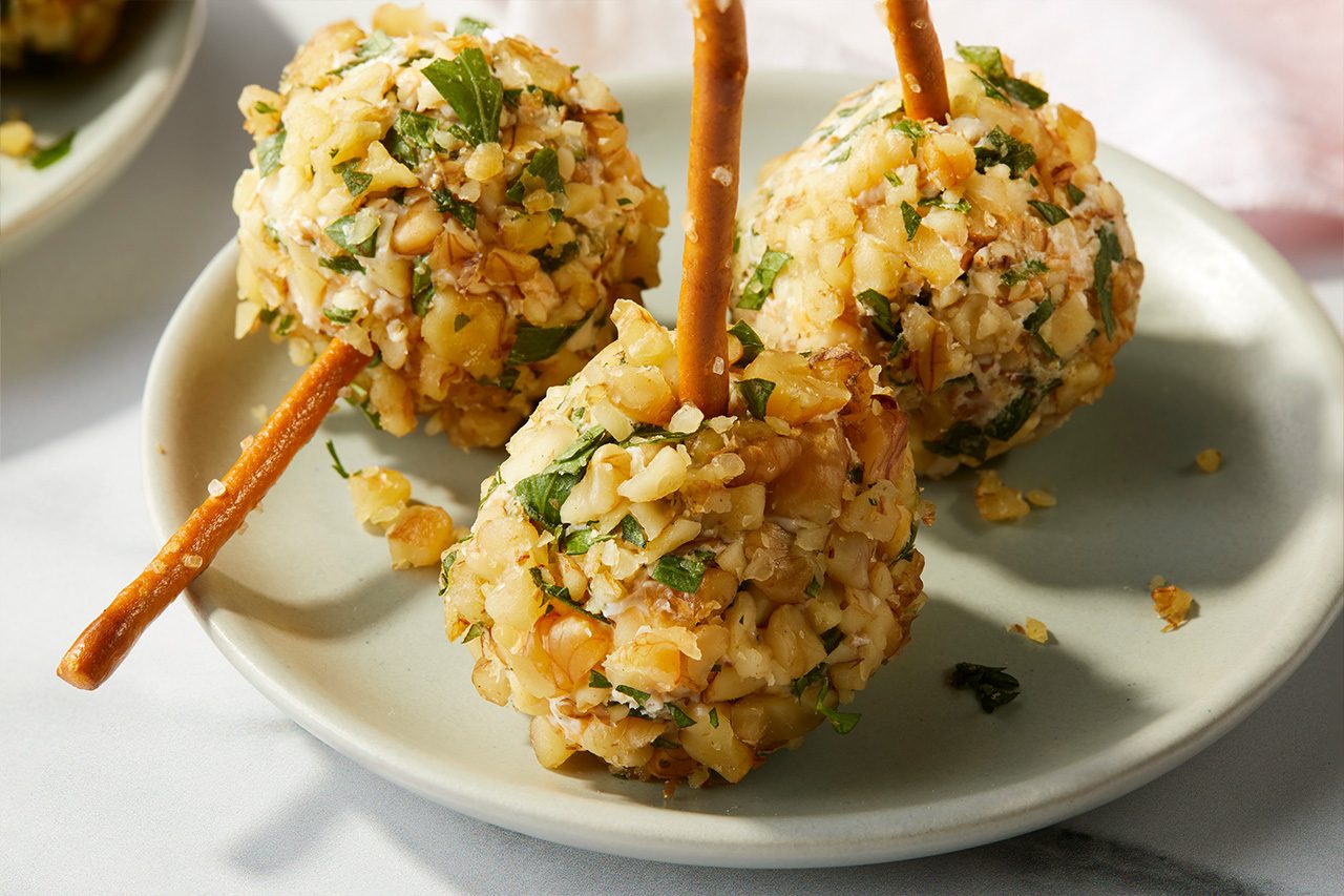 Eye Level horizontal image of three Dilly Mini Cheese Balls arranged on a light gray plate. Each mini cheese ball is coated with chopped walnuts and fresh parsley, and has a pretzel stick inserted for serving. The appetizers are presented against a soft, light background. | [Original] RMS 38691 Mini Dill Pickle Cheese Balls; Horizontal Push