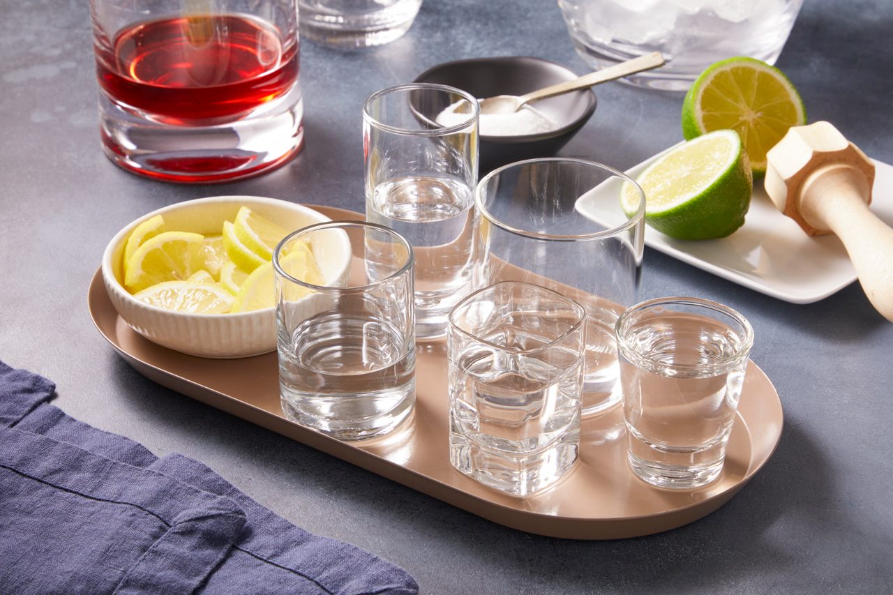 Multiple shot glasses containing clear spirits and lime juice are arranged, alongside bowls of sugar and lemon slices, with cranberry juice, ice cubes, and halved limes.