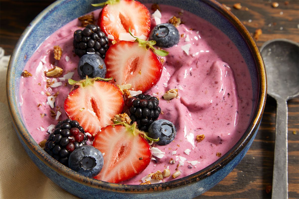 3/4 angle view shot of a bowl of no-churn frozen yogurt with a creamy berry swirl, topped with fresh sliced strawberries, blueberries, blackberries, shredded coconut, and chopped pistachios, served on a rustic wooden surface with a spoon alongside