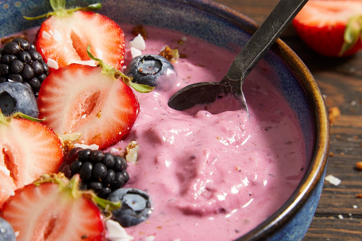 Close-up shot of a spoon scooping creamy no-churn frozen yogurt with a berry swirl, garnished with fresh strawberries, blueberries, blackberries, shredded coconut, and chopped pistachios in a rustic ceramic bowl