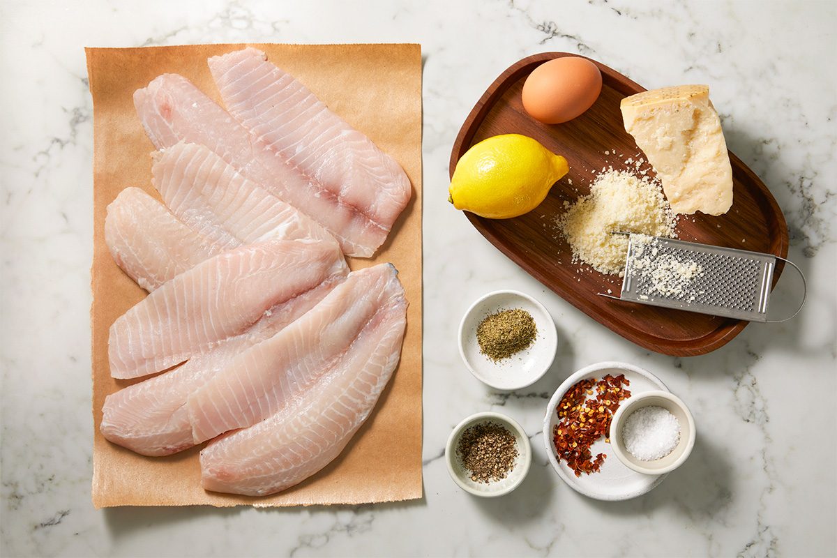A horizontal overhead image shows raw tilapia fillets arranged on parchment alongside a lemon, egg, grated Parmesan cheese, spices, and a grater on a marble surface.