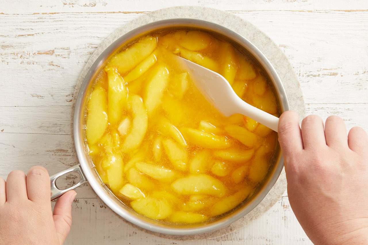 Overhead shot of a saucepan filled with sliced peaches simmering in a glossy syrup, as hands stir the mixture with a white spatula on a light wooden surface;