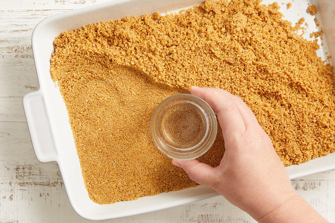 Overhead shot of a baking dish filled with an even layer of graham cracker crumb mixture as a hand sprinkles ground cinnamon from a small glass bowl over the surface;