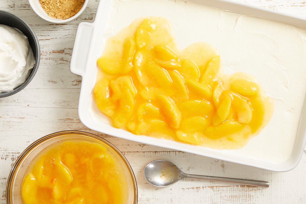 Overhead shot of sliced peaches spread evenly in a white baking dish over a creamy filling layer, with extra peach filling, graham cracker crumbs, and the remaining cream mixture arranged nearby on a light wooden surface;