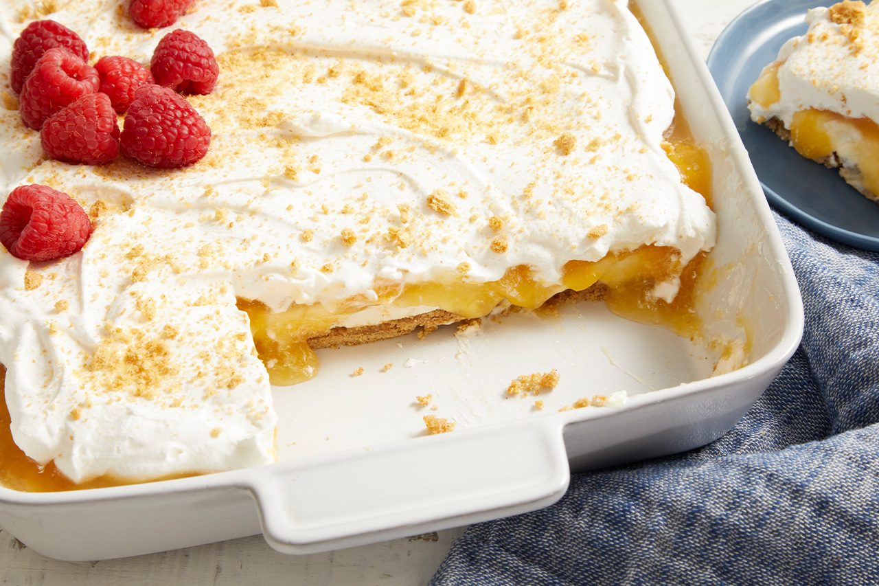 3/4 angle view shot of Easy Peach Delight in a white baking dish with a serving removed, revealing layers of graham cracker crust, creamy filling, and sweet peaches beneath a fluffy whipped topping. Fresh raspberries and graham cracker crumbs garnish the dessert, with a plated serving visible in the background;