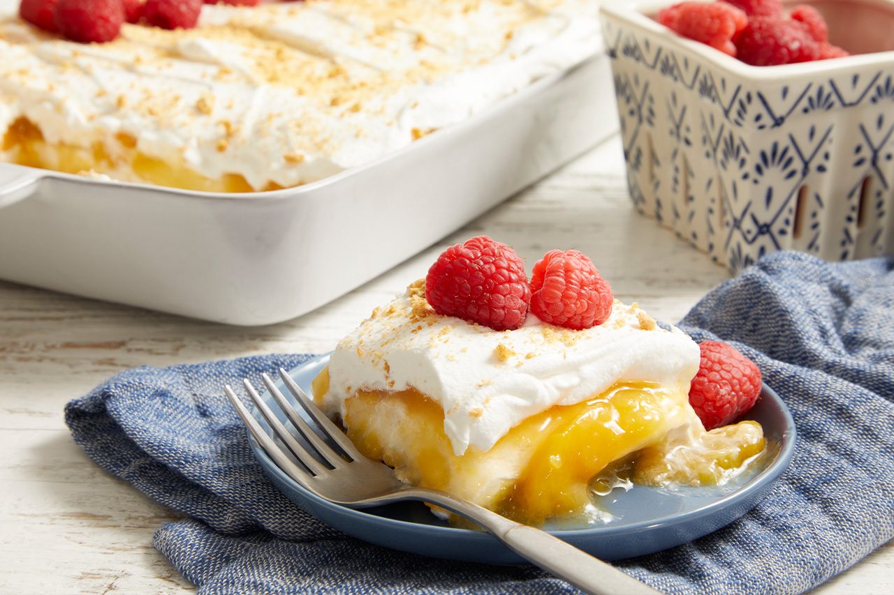 3/4 angle view shot of a plated serving of Easy Peach Delight topped with fluffy whipped topping, fresh raspberries, and graham cracker crumbs, with the remaining dessert in a baking dish blurred in the background on a light wooden surface;