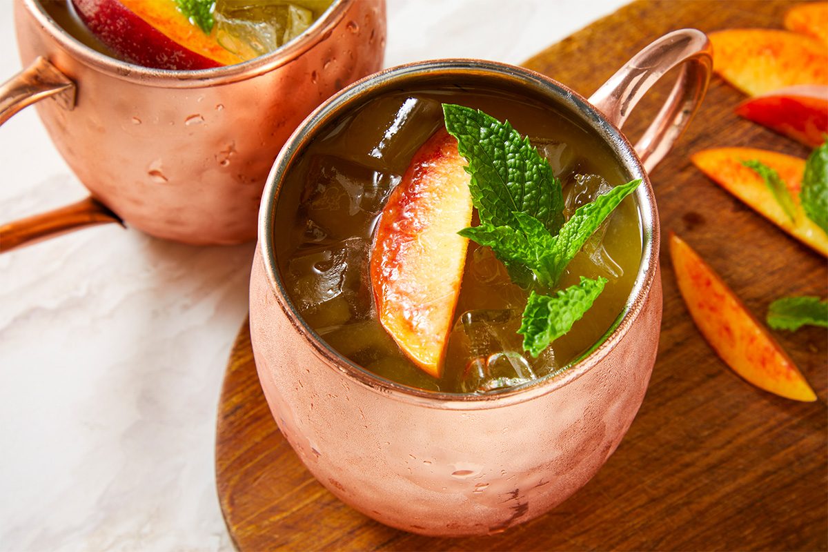 Eye Level horizontal image of two copper mugs filled with an iced drink, garnished with a peach slice and fresh mint sprigs. Additional peach slices and mint leaves are scattered on a wooden board next to the mugs, set against a white marble-like surface.
