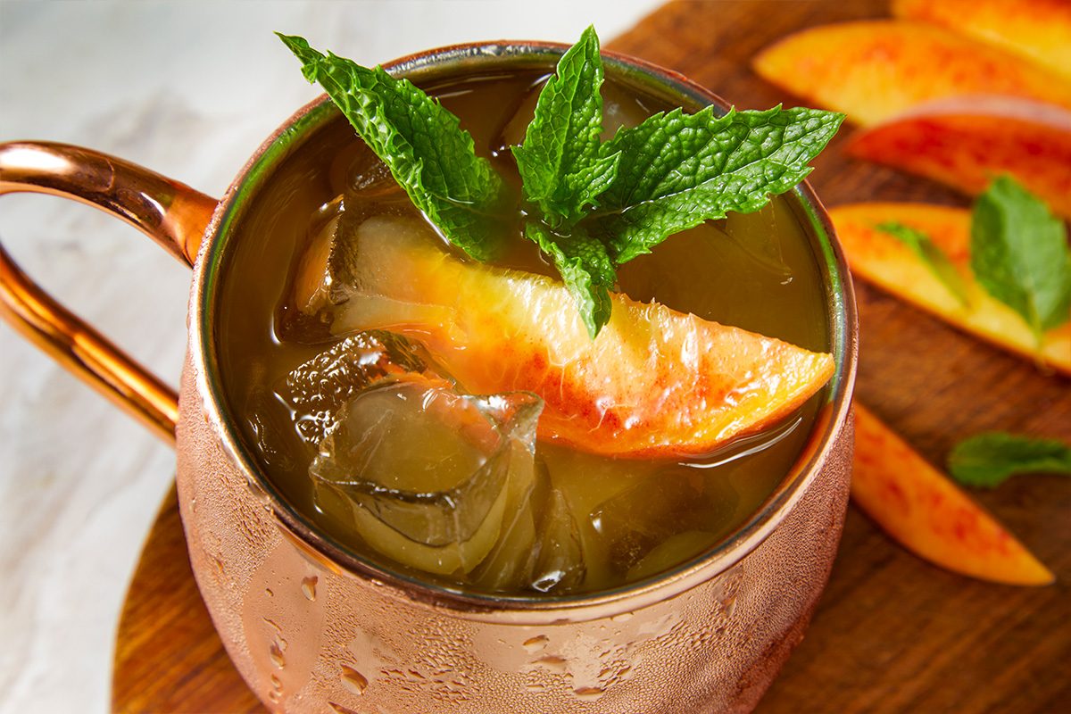 Overhead horizontal image of a cold beverage served in a copper mug. The drink contains ice cubes, a peach slice, and a fresh mint sprig garnish. Additional peach slices are visible on a wooden surface in the background.