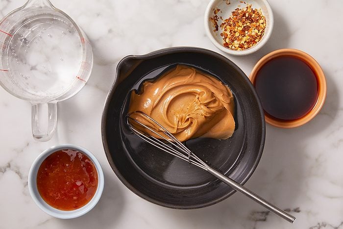 A bowl of creamy peanut butter with a whisk, surrounded by small bowls of chili flakes, soy sauce, water, and sweet chili sauce on a marble surface.