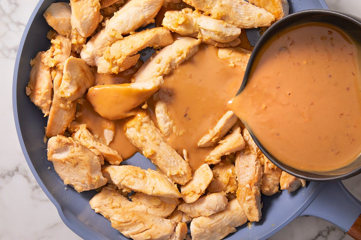 Sliced cooked chicken breast in a bowl with creamy brown sauce being poured over it, all on a light-colored surface.