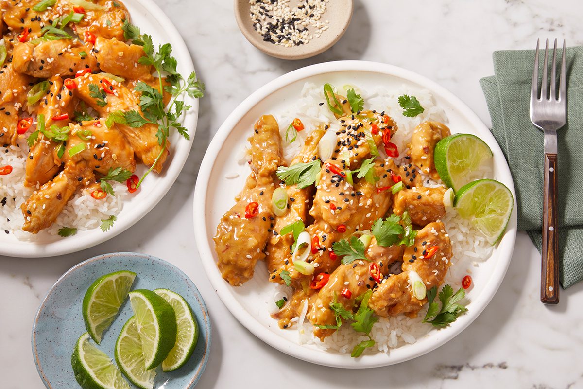Plates of crispy chicken topped with sesame seeds, sliced chili, and cilantro, served over rice with lime wedges on the side. A fork, napkin, and small bowls of lime and sesame seeds are nearby.