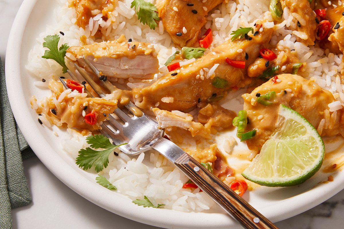 A plate of white rice topped with creamy chicken curry, sliced red chilies, black sesame seeds, cilantro, and a lime wedge. A fork rests on the plate.