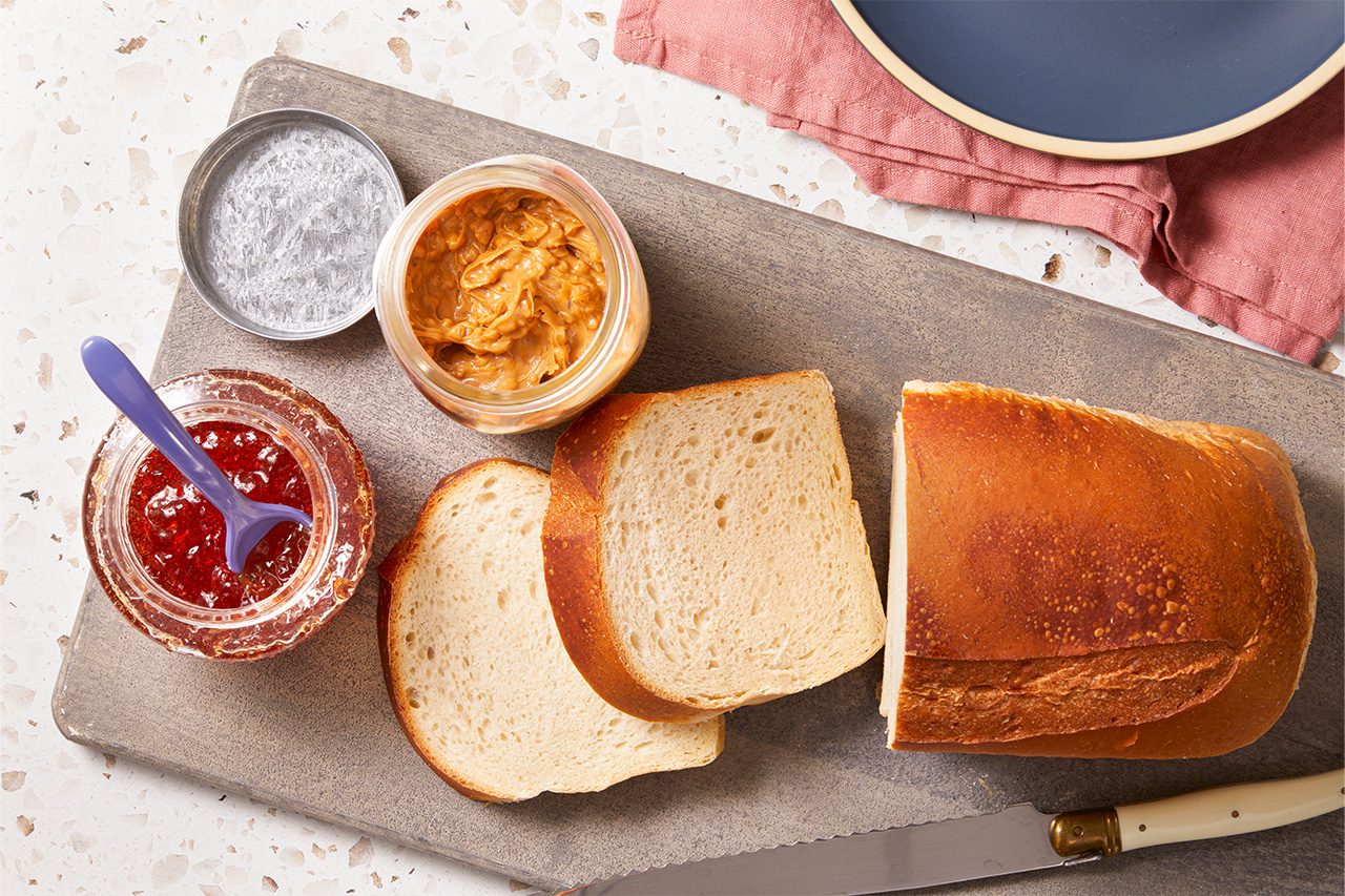 Overhead horizontal image of ingredients for a peanut butter and jelly sandwich. A partially sliced loaf of white bread, two bread slices, an open jar of chunky peanut butter, and an open jar of red jelly with a spoon are arranged on a wooden cutting board.
