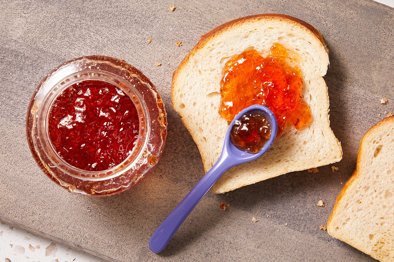 Overhead horizontal image of a slice of white bread topped with a dollop of orange marmalade. A purple spoon holding red jam rests on the bread, next to an open jar of red jam, all arranged on a dark cutting board.