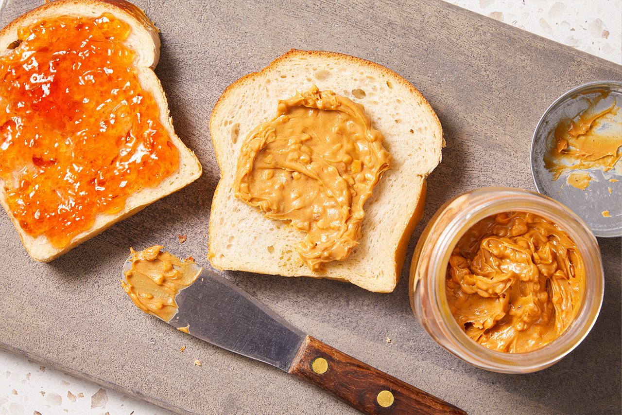 Overhead horizontal image of ingredients for a peanut butter and jelly sandwich. One slice of white bread is spread with orange jam, while another slice features chunky peanut butter. An open jar of chunky peanut butter, its lid, and a knife with peanut butter on the blade are also shown.