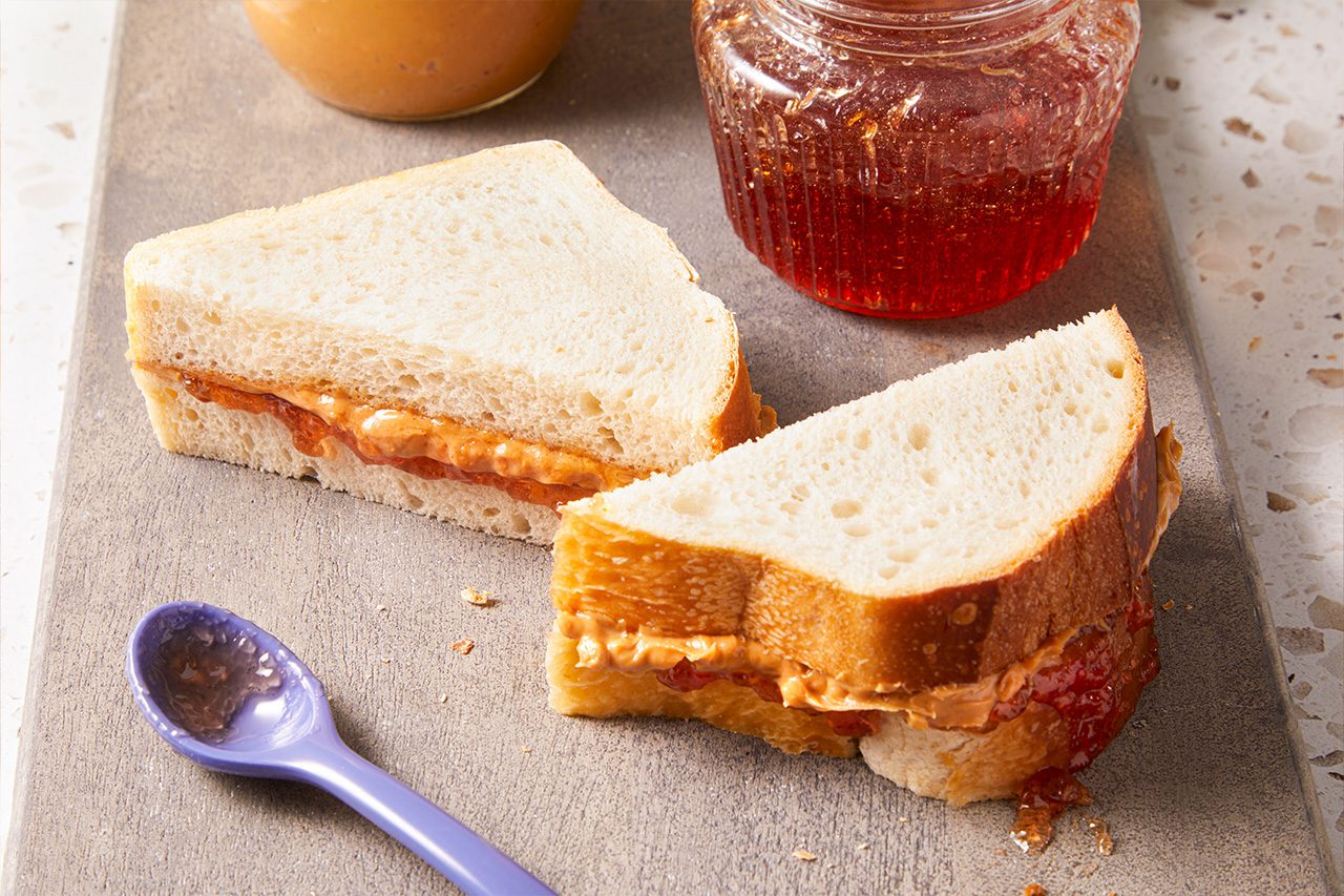 Overhead horizontal image of a peanut butter and jelly sandwich cut diagonally into two halves, resting on a gray cutting board. A jar of peanut butter and a jar of jelly are visible in the background, along with a purple spoon holding a dollop of jelly in the foreground. The sandwich filling clearly shows creamy peanut butter and fruit jelly between slices of white bread.