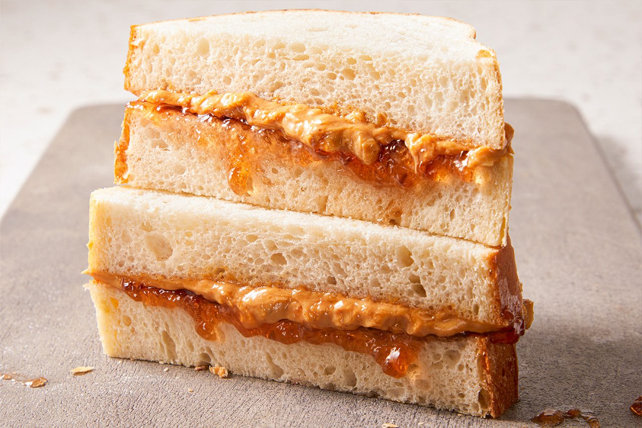 Eye Level horizontal image of a peanut butter and jelly sandwich. The sandwich is cut in half and stacked, showcasing the layers of creamy peanut butter and sweet fruit jelly between two slices of white bread. Some jelly and peanut butter are visible oozing slightly from the sides.
