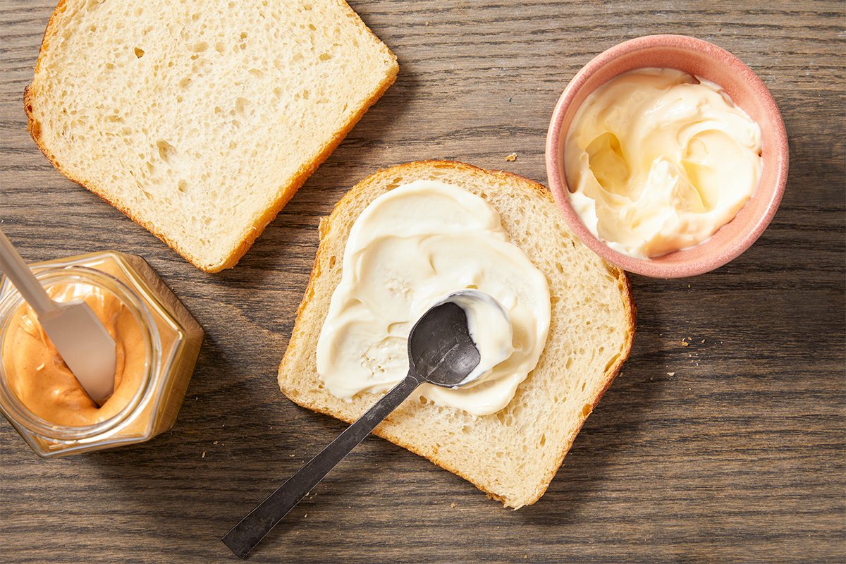 Overhead horizontal image of two slices of white bread placed on a wooden cutting board. Alongside the bread are a hexagonal jar of peanut butter with a knife coated in peanut butter, and a pink bowl containing mayonnaise with a spoon.