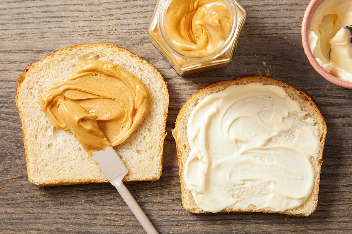 Overhead horizontal image of two slices of bread on a wooden surface. One slice has peanut butter being spread with a spatula, while the other is covered with mayonnaise. An open jar of peanut butter and a small bowl of mayonnaise with a spoon are also visible.