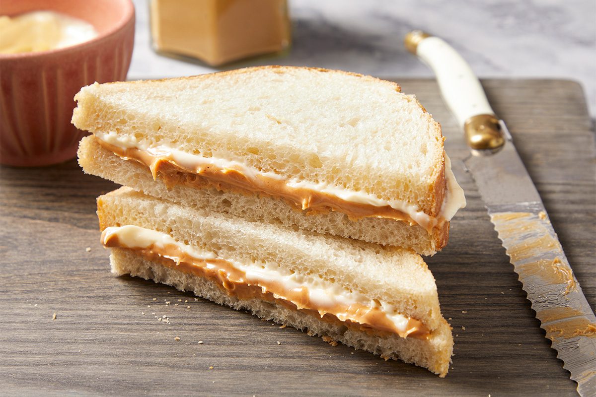 Eye Level horizontal image of a peanut butter and mayonnaise sandwich cut in half and stacked on a wooden cutting board. A knife with peanut butter residue rests beside the sandwich, with a pink bowl of mayonnaise and a jar of peanut butter blurred in the background.