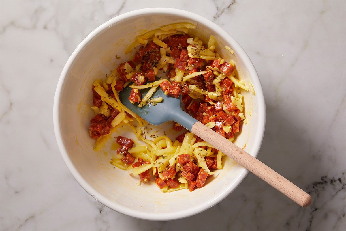 Overhead horizontal image of a mixing bowl filled with diced pepperoni, shredded mozzarella cheese, and dried oregano being combined with a spatula. This image captures the flavor-building stage of the pepperoni pinwheels recipe.
