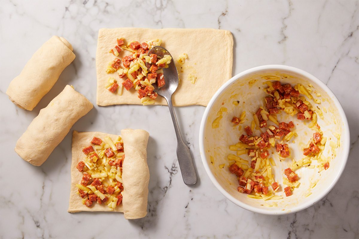 Overhead horizontal image showing crescent roll dough rectangles topped with pepperoni filling and partially rolled into logs. The scene illustrates the assembly process before slicing.